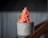 A creamy, pink sorbet is served in a simple, textured gray cup. The sorbet is garnished with a thin round slice of dried lime and small zest pieces, adding a touch of green color. The background features a blurred, corrugated texture.