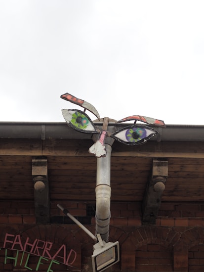 A unique and creative sculpture resembling a face is made from a combination of industrial and recycled materials, including a metal pipe and painted elements forming eyes and lips. It is mounted on a brick wall, with some graffiti partially visible. The background includes a wooden roof structure.