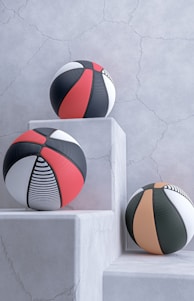 Three basketballs with modern designs are placed on white, textured stone pedestals against a cracked, light gray wall. The balls feature patterns in black, red, white, and orange, including stripes and solid panels.