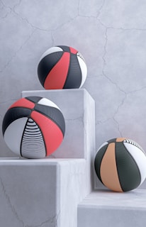 Three basketballs with modern designs are placed on white, textured stone pedestals against a cracked, light gray wall. The balls feature patterns in black, red, white, and orange, including stripes and solid panels.