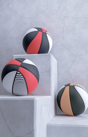 Three basketballs with modern designs are placed on white, textured stone pedestals against a cracked, light gray wall. The balls feature patterns in black, red, white, and orange, including stripes and solid panels.