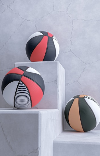Three basketballs with modern designs are placed on white, textured stone pedestals against a cracked, light gray wall. The balls feature patterns in black, red, white, and orange, including stripes and solid panels.