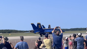 A group of people is gathered near a red safety barrier, observing and taking photos of two navy blue and yellow fighter jets parked on a runway. The sky is clear, and there are trees in the background.