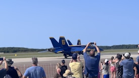 A group of people is gathered near a red safety barrier, observing and taking photos of two navy blue and yellow fighter jets parked on a runway. The sky is clear, and there are trees in the background.