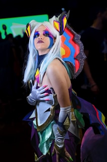A person in a vibrant and colorful costume with exaggerated makeup and a white wig. The outfit features bright geometric shapes in hues of purple, blue, orange, and yellow, and includes large, ornate shoulder pieces. The background appears blurred and dark, highlighting the elaborate attire.