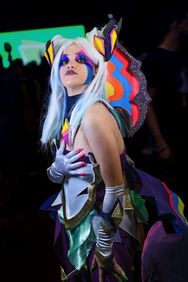 A person in a vibrant and colorful costume with exaggerated makeup and a white wig. The outfit features bright geometric shapes in hues of purple, blue, orange, and yellow, and includes large, ornate shoulder pieces. The background appears blurred and dark, highlighting the elaborate attire.