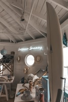 A cozy, seaside-themed interior with nautical decor, including a surfboard, hats, and a neon sign reading 'California Dreaming'. The room has a small, round window and is adorned with light wooden accents and maritime elements, such as beach chairs and striped fabrics.