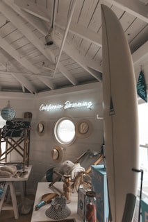 A cozy, seaside-themed interior with nautical decor, including a surfboard, hats, and a neon sign reading 'California Dreaming'. The room has a small, round window and is adorned with light wooden accents and maritime elements, such as beach chairs and striped fabrics.