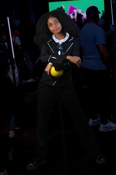 A person wearing a black suit with a white collar and holding a yellow ball stands with arms crossed. The background includes a crowd of people and a bright screen displaying colorful graphics.