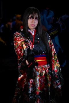 A person is dressed in a vibrant, traditional Japanese-inspired outfit with floral patterns and a striking red and black color scheme. The individual has long dark hair and is standing in a dimly lit environment, possibly at a cosplay event or gathering. The background is out of focus, with some other figures present.