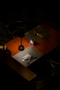 A dimly lit table setting is seen with a dark wooden surface and chairs. Two placemats are placed on the table, one labeled 'SIMPLE,' with black cutlery arranged on top. A small menu or promotional card with a QR code stands upright near the center of the table.