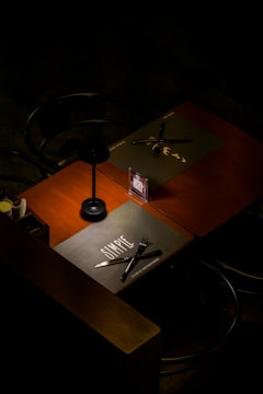 A dimly lit table setting is seen with a dark wooden surface and chairs. Two placemats are placed on the table, one labeled 'SIMPLE,' with black cutlery arranged on top. A small menu or promotional card with a QR code stands upright near the center of the table.
