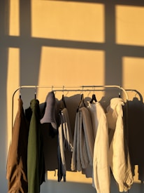A row of various colored clothes hanging on a rack, with sunlight casting shadows on the wall behind. The garments include jackets, sweaters, and hoodies in neutral and earthy tones.