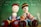 Two crocheted dolls sit in front of a green chalkboard with white chalk scribbles. The dolls are adorned in colorful striped outfits with large button accents as shoes. Their faces are simplistic with stitched features.