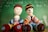 Two crocheted dolls sit in front of a green chalkboard with white chalk scribbles. The dolls are adorned in colorful striped outfits with large button accents as shoes. Their faces are simplistic with stitched features.