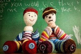 Two crocheted dolls sit in front of a green chalkboard with white chalk scribbles. The dolls are adorned in colorful striped outfits with large button accents as shoes. Their faces are simplistic with stitched features.