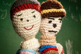 Two crocheted dolls are positioned closely, one with a smiling face and a red beanie, while the other has a more serious expression with a knitted hat featuring a yellow stripe. They are set against a blurred green background resembling a chalkboard.