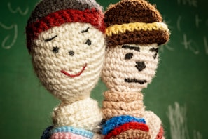 Two crocheted dolls are positioned closely, one with a smiling face and a red beanie, while the other has a more serious expression with a knitted hat featuring a yellow stripe. They are set against a blurred green background resembling a chalkboard.