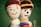 Two crocheted dolls are positioned closely, one with a smiling face and a red beanie, while the other has a more serious expression with a knitted hat featuring a yellow stripe. They are set against a blurred green background resembling a chalkboard.