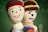 Two crocheted dolls are positioned closely, one with a smiling face and a red beanie, while the other has a more serious expression with a knitted hat featuring a yellow stripe. They are set against a blurred green background resembling a chalkboard.