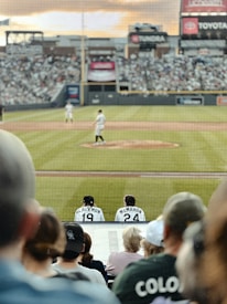 A baseball game scene from the perspective of the audience behind home plate. The field is visible with players on the pitcher’s mound and the field. The focus is on two players seated with jersey numbers 19 and 24, named Blackmon and McMahon. The stadium is filled with spectators and advertisements are visible on the walls.