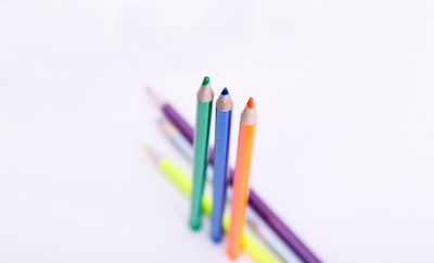 A collection of colorful pencils arranged on a white background, with three pencils standing upright in the foreground. The colors include green, orange, and blue, while other pencils lie flat in the background.
