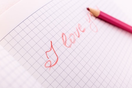 A piece of graph paper features the handwritten phrase 'I love you' in red cursive. A red colored pencil is placed next to the writing, indicating it was used to write the message.