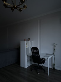 A minimalist home office features a sleek white desk paired with a black office chair. On the desk sits a closed laptop. Next to the desk is a white shelving unit containing a few books and decorative items. A small potted plant is placed on top of the shelf, while a tall plant stands on the floor nearby. The room is dimly lit, creating a calm and subdued atmosphere.