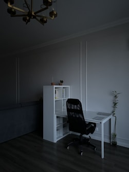 A minimalist home office features a sleek white desk paired with a black office chair. On the desk sits a closed laptop. Next to the desk is a white shelving unit containing a few books and decorative items. A small potted plant is placed on top of the shelf, while a tall plant stands on the floor nearby. The room is dimly lit, creating a calm and subdued atmosphere.