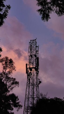 A communication tower stands tall against a backdrop of a vibrant sunset sky, surrounded by silhouettes of trees. The sky is rich with colors, blending different shades of pink, purple, and orange. The tower's structure is prominent, capturing the evening's serene and peaceful ambiance.