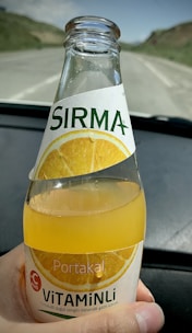 A hand holding a glass bottle of orange-flavored carbonated mineral drink. The bottle features a label with an image of an orange slice and text indicating vitamin content. In the background, there is an out-of-focus view of a road and landscape.