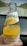 A hand holding a glass bottle of orange-flavored carbonated mineral drink. The bottle features a label with an image of an orange slice and text indicating vitamin content. In the background, there is an out-of-focus view of a road and landscape.