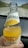 A hand holding a glass bottle of orange-flavored carbonated mineral drink. The bottle features a label with an image of an orange slice and text indicating vitamin content. In the background, there is an out-of-focus view of a road and landscape.