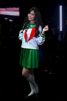 A person dressed in a green and white school uniform with a red tie, standing confidently with a slight smile. The outfit includes a pleated green skirt, white knee-high socks, and dark shoes. The background is dimly lit, with a bright vertical light source to the side.