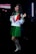 A person dressed in a green and white school uniform with a red tie, standing confidently with a slight smile. The outfit includes a pleated green skirt, white knee-high socks, and dark shoes. The background is dimly lit, with a bright vertical light source to the side.