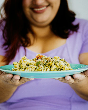 A smiling woman holding a colorful plate of healthy food in a bright dining room.