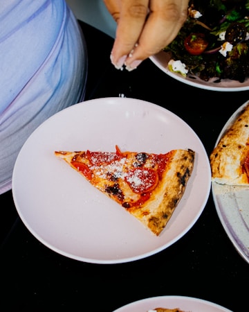 A hand sprinkles Parmesan cheese over a slice of pepperoni pizza on a white plate. Nearby, there is a salad with cherry tomatoes and greens, partially visible. Another pizza on a silver tray can be seen at the edge.