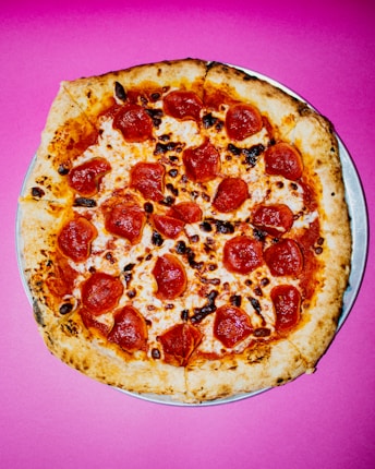 A round pepperoni pizza with a crispy crust is placed on a vibrant pink background. The pizza is topped with melted cheese and evenly distributed slices of pepperoni, some of which have a slightly charred appearance.