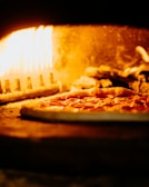 A pizza with pepperoni toppings is being baked in a wood-fired oven. The flames are visible and illuminate the interior of the oven with a warm, orange glow.