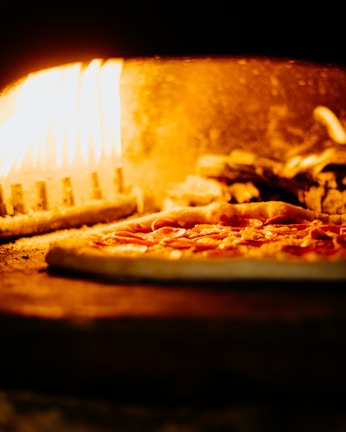 A pizza with pepperoni toppings is being baked in a wood-fired oven. The flames are visible and illuminate the interior of the oven with a warm, orange glow.