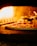 A pizza with pepperoni toppings is being baked in a wood-fired oven. The flames are visible and illuminate the interior of the oven with a warm, orange glow.