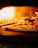 A pizza with pepperoni toppings is being baked in a wood-fired oven. The flames are visible and illuminate the interior of the oven with a warm, orange glow.