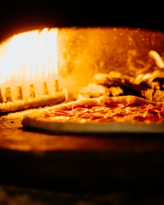 A pizza with pepperoni toppings is being baked in a wood-fired oven. The flames are visible and illuminate the interior of the oven with a warm, orange glow.
