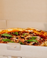 A freshly-baked pizza with a golden-brown crust is topped with melted cheese, tomato sauce, and fresh basil leaves. It is placed inside an open cardboard pizza box.