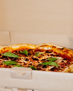 A freshly-baked pizza with a golden-brown crust is topped with melted cheese, tomato sauce, and fresh basil leaves. It is placed inside an open cardboard pizza box.