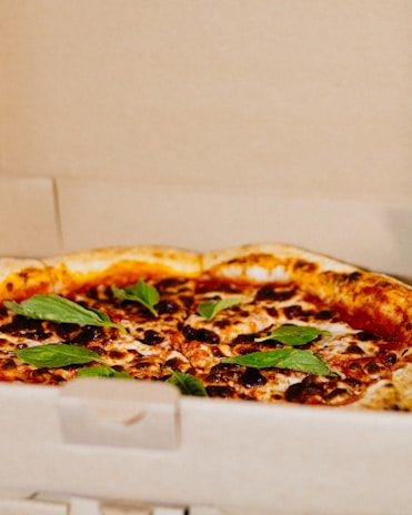 A freshly-baked pizza with a golden-brown crust is topped with melted cheese, tomato sauce, and fresh basil leaves. It is placed inside an open cardboard pizza box.