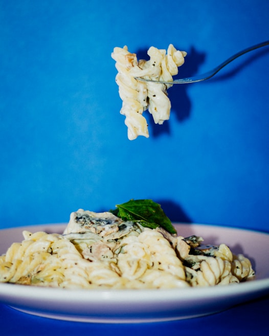 A plate of creamy pasta garnished with fresh basil sits against a vibrant blue background, with a fork holding up a bite of the pasta.