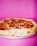 A pepperoni pizza with a thick golden crust is placed against a vibrant pink background. The pizza is topped with melted cheese and slices of pepperoni, with some slight charring visible on the crust.