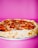 A pepperoni pizza with a thick golden crust is placed against a vibrant pink background. The pizza is topped with melted cheese and slices of pepperoni, with some slight charring visible on the crust.
