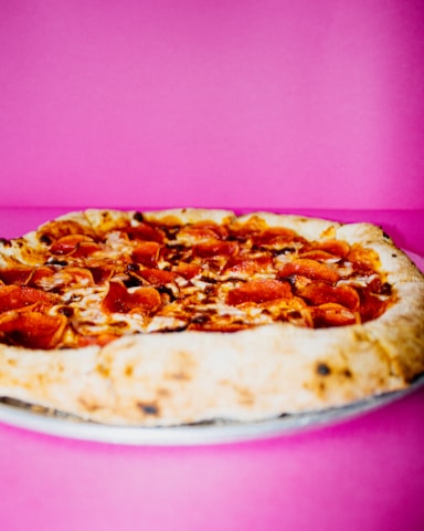 A pepperoni pizza with a thick golden crust is placed against a vibrant pink background. The pizza is topped with melted cheese and slices of pepperoni, with some slight charring visible on the crust.